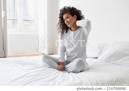 A young Hispanic woman sits on a white bed in a well-lit bedroom, dressed in comfortable clothing, holding her neck with a pained expression that suggests discomfort or neck pain A young Hispanic woman sits on a white bed in a well-lit bedroom, dressed in comfortable clothing, holding her neck with a pained expression that suggests discomfort or neck pain 114708690