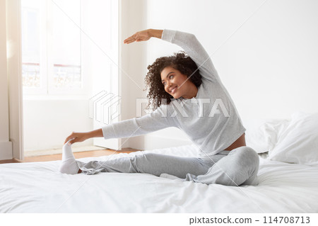 Hispanic woman is stretching her body while lying on a bed with white sheets. She is extending her arms and legs to loosen up her muscles after waking up. 114708713