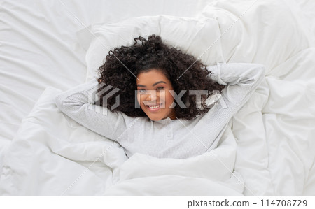 Hispanic woman is laying peacefully in a bed with a white comforter. She appears relaxed and comfortable, surrounded by the soft and fluffy bedding, smiling at camera 114708729