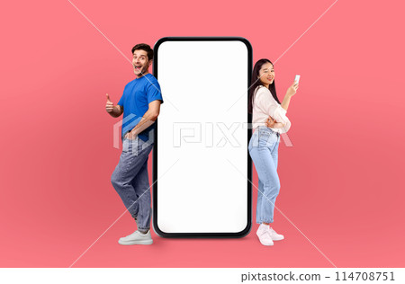 Multiethnic lovers stand on either side of a large smartphone, both expressing excitement and approval with thumbs up gestures Multiethnic lovers stand on either side of a large smartphone, both expressing excitement and approval with thumbs up gestures 114708751