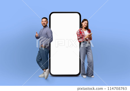 Asian couple standing next to a large-sized telephone, looking at the device with curiosity. The phone appears oversized in comparison to the people, creating a unique visual contrast. 114708763