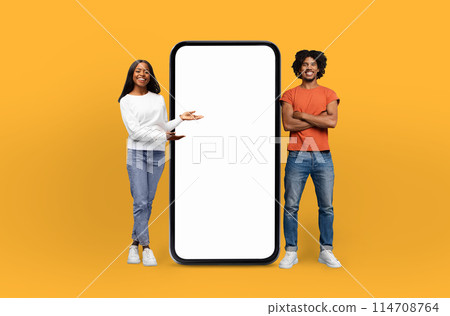 A smiling African American couple casually presents the blank screen of an oversized smartphone mockup, indicating its features or potential content 114708764