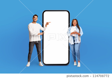 Multiethnic man and woman are standing side by side next to large phone, looking at the screen and appearing to have conversation. The phone is significantly bigger than them, emphasizing its size. Multiethnic man and woman are standing side by side next to large phone, looking at the screen and appearing to have conversation. The phone is significantly bigger than them, emphasizing its size. 114708772