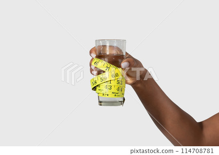 Cropped of black woman is shown holding a measuring tape up to a glass of water, possibly measuring the height or volume of the liquid, white background Cropped of black woman is shown holding a measuring tape up to a glass of water, possibly measuring the height or volume of the liquid, white background 114708781