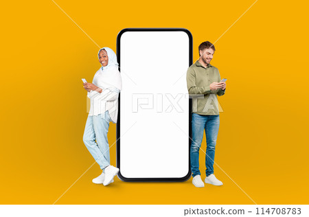 European man and black woman in hijab standing side by side, both looking at a cell phone in their hands, smartphone with white blank screen between them European man and black woman in hijab standing side by side, both looking at a cell phone in their hands, smartphone with white blank screen between them 114708783
