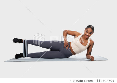 A cheerful African American woman in sportswear maintains a side plank pose with resistance band on her legs, engaging her core muscles A cheerful African American woman in sportswear maintains a side plank pose with resistance band on her legs, engaging her core muscles 114708795