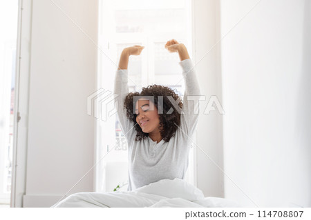 Hispanic woman is waking up in bed, stretching with her arms raised up in the air. She is lying down on a white bedsheet with pillows in the background. 114708807