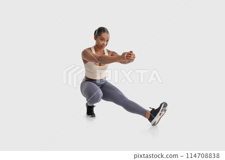 African American woman is shown doing a squat exercise on a plain white background. She is bending her knees and lowering her hips while keeping her back straight and arms extended in front of her 114708838