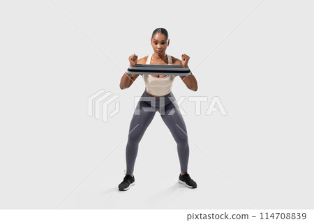A focused African American woman is engaging in a fitness routine using a resistance band to strengthen her upper body muscles. She maintains a squat position, highlighting her athletic form 114708839