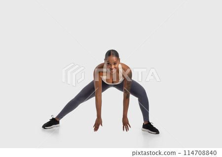 African American woman is seen performing a squat exercise on a white background. She is bending her knees while keeping her back straight, engaging her leg muscles. The white background 114708840