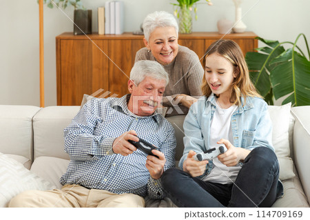 Happy family at home. Old senior man grandfather woman grandmother girl granddaughter playing video game with joysticks at home. Grandparent teenager two generations using gamepads for video game 114709169