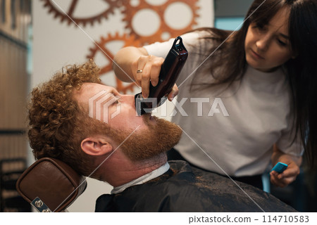 Red-haired bearded man sitting in barbershop and female barber making hairdo to guest Red-haired bearded man sitting in barbershop and female barber making hairdo to guest 114710583