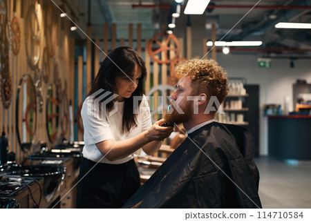 Red-haired bearded man sitting in barbershop and female barber making hairdo to guest Red-haired bearded man sitting in barbershop and female barber making hairdo to guest 114710584