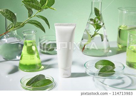 The sample tube of cosmetic without label displayed in center, petri dish with green tea leaves, an erlenmeyer of green tea extract and other experiment utensils in back. Mock up for advertising The sample tube of cosmetic without label displayed in center, petri dish with green tea leaves, an erlenmeyer of green tea extract and other experiment utensils in back. Mock up for advertising 114713983