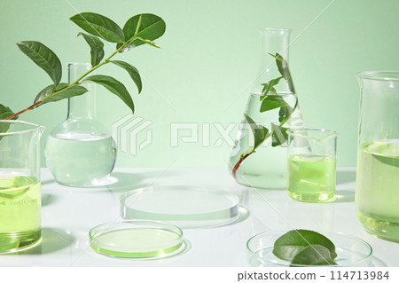Glassware in laboratory placed on white countertop against light green background, a empty round glass pedestal for showing product in center, plenty lab items decorated around. Space for design 114713984