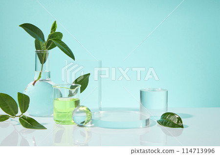 White counter top containing a few blank glass pedestal, a beaker with green fluid, crystal ball and a boiling flask filled by transparent liquid, green tea decoration. Front view, copy space 114713996