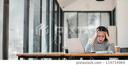 A woman is sitting at a desk with a laptop and looking very upset. She is surrounded by a window and a potted plant A woman is sitting at a desk with a laptop and looking very upset. She is surrounded by a window and a potted plant 114714969