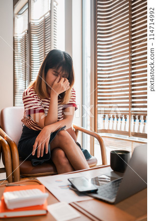 A woman is sitting in a chair with her head in her hands. She is wearing a striped shirt and a black skirt. The room has wooden blinds and a laptop on the table A woman is sitting in a chair with her head in her hands. She is wearing a striped shirt and a black skirt. The room has wooden blinds and a laptop on the table 114714992