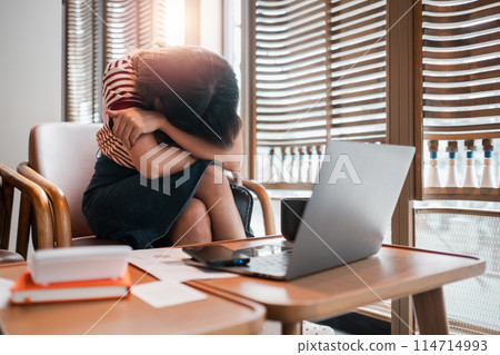 A woman is sitting in a chair with her head on her hands. She is crying and she is in distress. The room has a laptop and a few books on a table A woman is sitting in a chair with her head on her hands. She is crying and she is in distress. The room has a laptop and a few books on a table 114714993