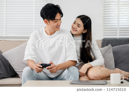 A man and woman are sitting on a couch playing a video game. The man is holding a controller and the woman is smiling. Scene is happy and relaxed 114715010