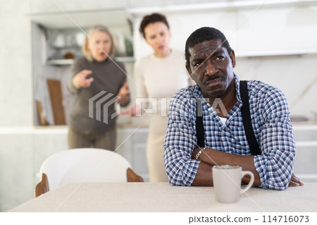 Middle-aged man sitting at the kitchen table with his back to women quarreling to him 114716073