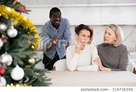 Domestic quarrel during Christmas celebration. Mom calms her daughter while her husband scolds her 114716394