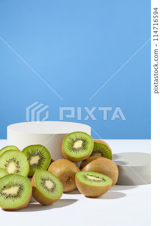 A cylinder podium in white color and a round podium in gray color decorated with a lot of kiwi cut in halves. Blue background. Kiwi fruit is well-known for its high vitamin C content 114716594