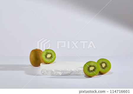 Against white background, a stone podium is decorated with some halves of kiwi fruit. Empty space for product presentation. Abstract composition in minimal design Against white background, a stone podium is decorated with some halves of kiwi fruit. Empty space for product presentation. Abstract composition in minimal design 114716601