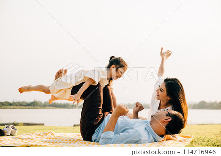 A joyful family moment in the park. Father holds his daughter up high as she smiles and flies like an airplane, while mother looks on with delight. Family fun at its best 114716946
