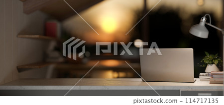 A laptop computer on a table in a room with dim lights from a lamp and sunset through the window. 114717135