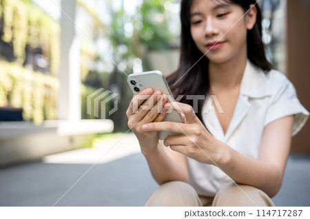 A beautiful Asian woman sits on the stairs outside the building and uses her smartphone. A beautiful Asian woman sits on the stairs outside the building and uses her smartphone. 114717287