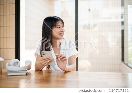 A smiling Asian woman gazing at the view outside while sitting indoors with a smartphone in her hand A smiling Asian woman gazing at the view outside while sitting indoors with a smartphone in her hand 114717351