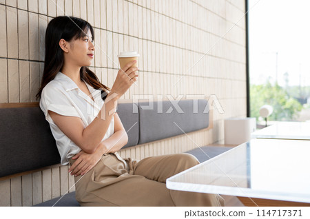 A beautiful Asian woman is gazing at the view outside while enjoying a coffee in the coffee shop. A beautiful Asian woman is gazing at the view outside while enjoying a coffee in the coffee shop. 114717371