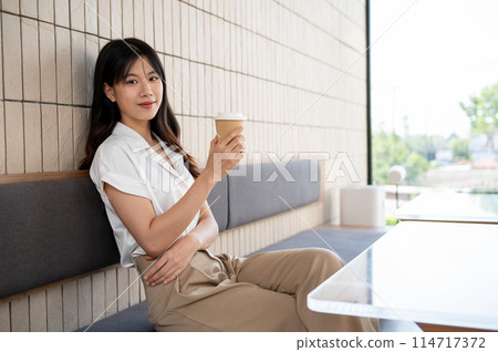 An attractive Asian woman sits in a coffee shop, a coffee cup in her hand, and smiles at the camera. An attractive Asian woman sits in a coffee shop, a coffee cup in her hand, and smiles at the camera. 114717372