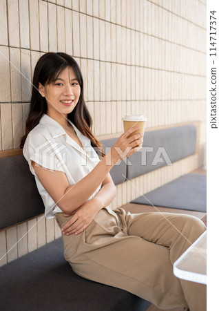 An attractive Asian woman sits in a coffee shop, a coffee cup in her hand, and smiles at the camera. An attractive Asian woman sits in a coffee shop, a coffee cup in her hand, and smiles at the camera. 114717374
