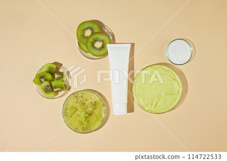 Top view photo at a white cosmetic tube without label featured in center with some round glass dish containing kiwi scrub and and fresh kiwi around. Mock up for designing with space for text 114722533