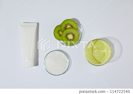 Photo for advertising with blank label tube in white color lay flat on white texture next to a dish of fresh kiwi and two glass dishes contain exfoliating cream in white and yellow. Space for text 114722540