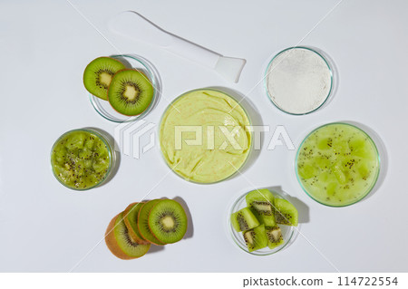 A dish containing yellow exfoliating scrub placed in center, surrounded by some dish of fresh kiwi over white texture. Photo from above view with empty space for advertising 114722554