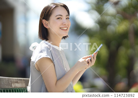 年輕女子坐在城市的長凳上，看著她的智慧型手機 114723892