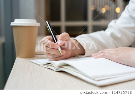 A cropped image of a woman sits in a coffee shop, writing something in a book or keeping her diary. 114725758