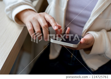 A close-up image of a woman in casual wear using her smartphone at a table indoors. 114725761