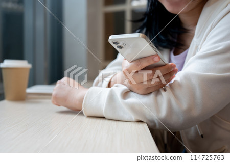 A cropped image of an Asian woman in casual clothes sits in a coffee shop, holds her smartphone. A cropped image of an Asian woman in casual clothes sits in a coffee shop, holds her smartphone. 114725763