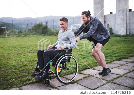 Young man in wheelchair spending free time outdoor with best friend, having fun in city. Male friendship. 114726216
