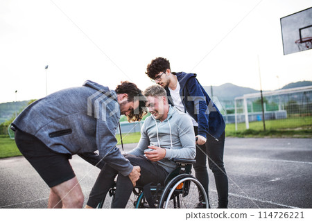 Young man in wheelchair spending free time outdoor with friends. Friends helping disabled man to sit in wheelchair. Male friendship. Young man in wheelchair spending free time outdoor with friends. Friends helping disabled man to sit in wheelchair. Male friendship. 114726221