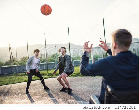 Disabled young man in a wheelchair playing basketball with his friends. Teamwrok and male friendship. 114726291