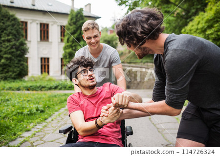 Young man in wheelchair spending free time outdoor with friends, having fun in city. Male friendship. Young man in wheelchair spending free time outdoor with friends, having fun in city. Male friendship. 114726324