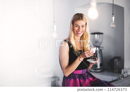 Beautiful blond woman holding cup of coffe, looking at camera. Female coffee shop business owner. 114726373