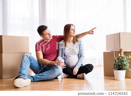 Beautiful young couple expecting a baby, moved into new home. Sitting among cardboard boxes. New beginnings. Beautiful young couple expecting a baby, moved into new home. Sitting among cardboard boxes. New beginnings. 114726581