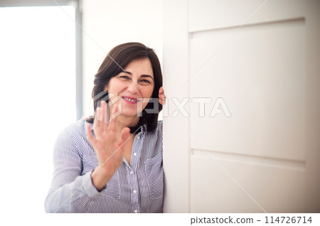 Beautiful mature woman waving, opening door to guest. Looking at camera and smiling, welcome. Beautiful mature woman waving, opening door to guest. Looking at camera and smiling, welcome. 114726714