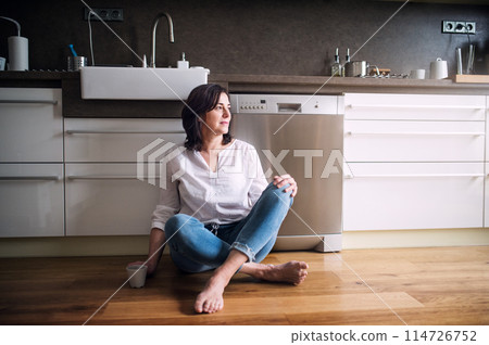 Beautiful mature woman sitting on kitchen floor, drinking coffee. Older woman living alone, enjoying peaceful weekend day Beautiful mature woman sitting on kitchen floor, drinking coffee. Older woman living alone, enjoying peaceful weekend day 114726752